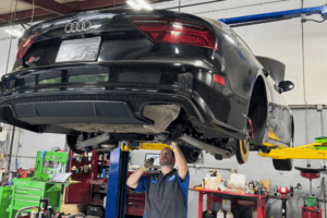 A technician performs an inspection on the underside of a black Audi S7, raised on a hydraulic lift in a professional auto repair shop. AutoImports of Denver is your BMW and Audi repair specialist for European auto repair in Denver, CO.